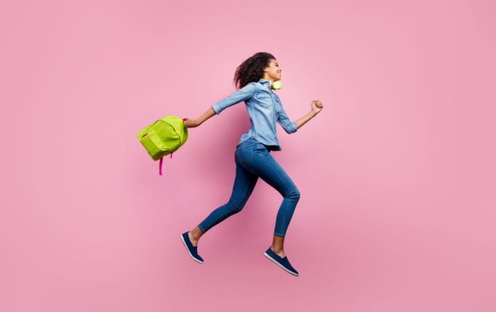 Woman in jeans and a shirt running with backpack in hand on pink background