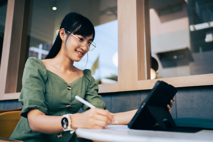 A smiling young woman writing down notes and looking at a tablet